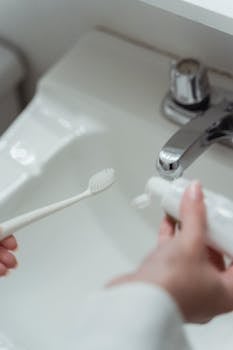 Close-up of hands holding a toothbrush and toothpaste over a bathroom sink.