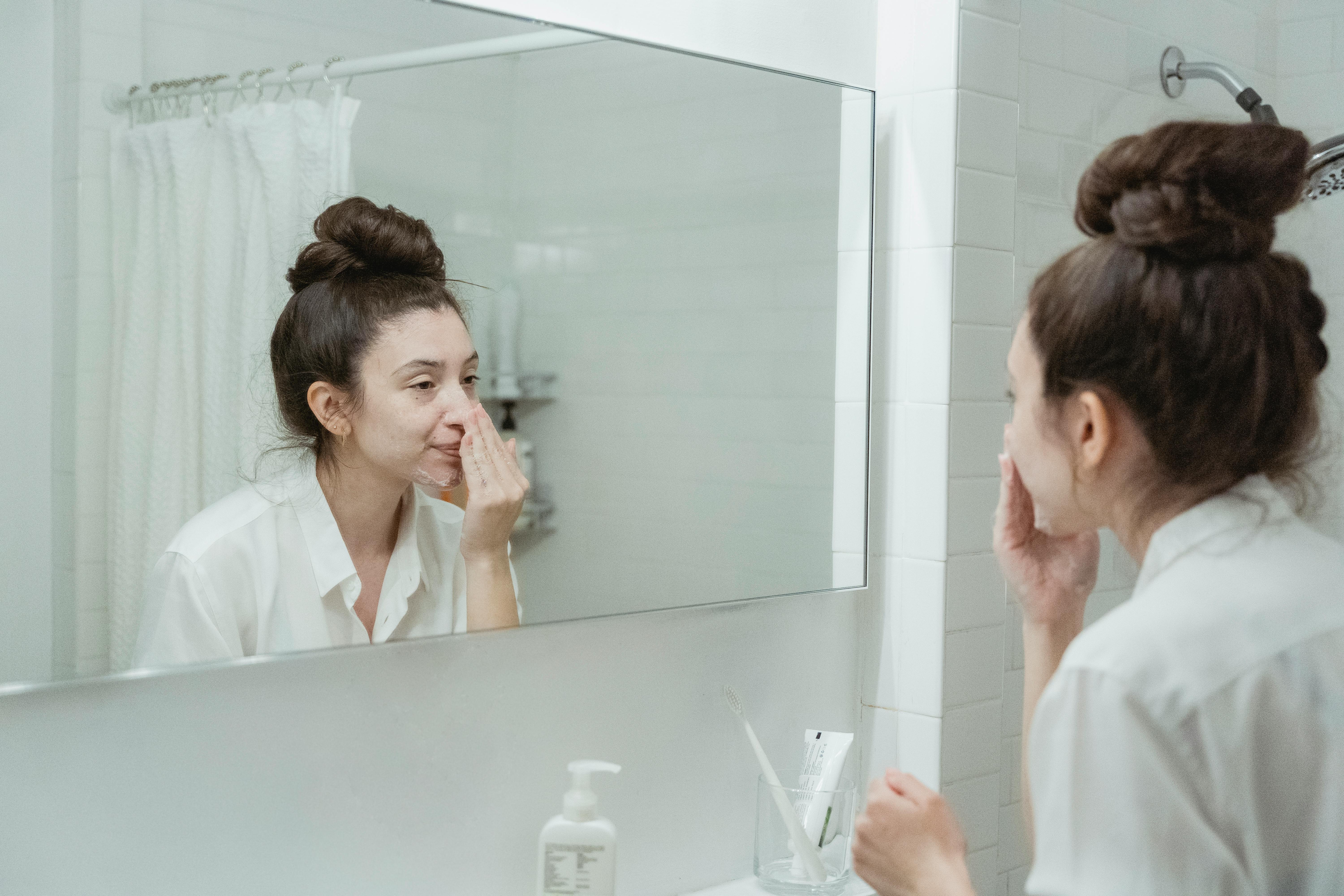 woman in a white bathroom washing face and looking mirror