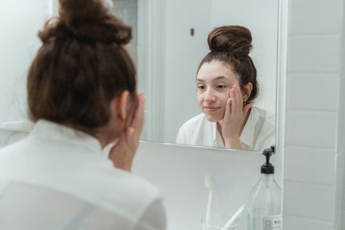 Free Woman looking at her reflection in the mirror while applying skincare products. Stock Photo