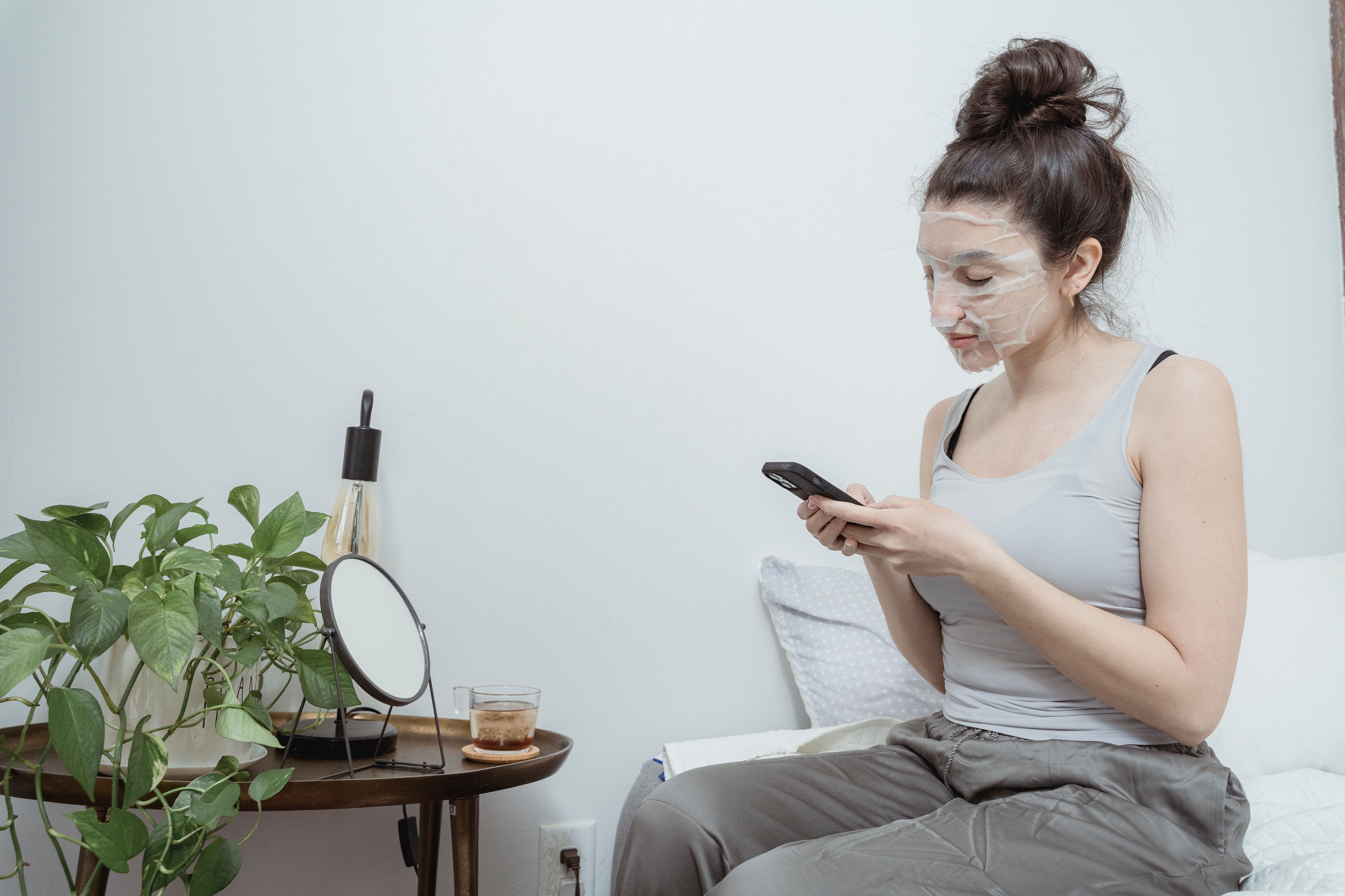 A woman with a facial sheet mask uses her smartphone while relaxing indoors.