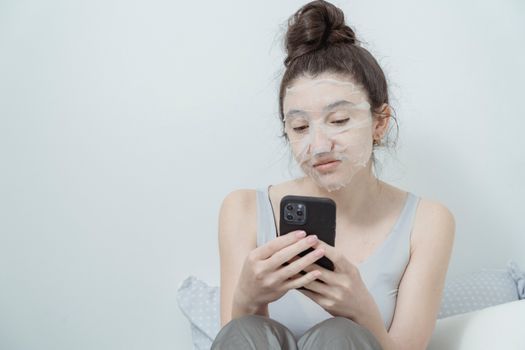 Young woman with a beauty mask sits indoors, browsing her smartphone, enjoying a skincare routine.