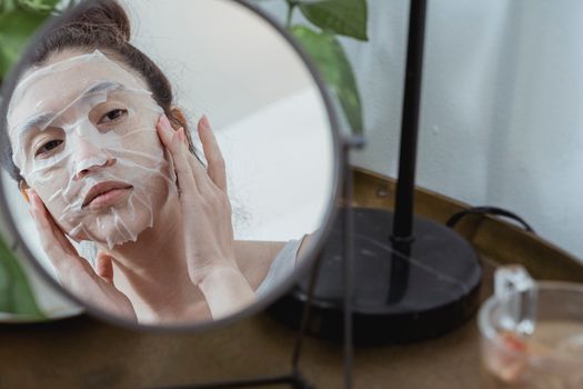A woman applies a cosmetic sheet mask while looking at her reflection in a mirror for skincare.