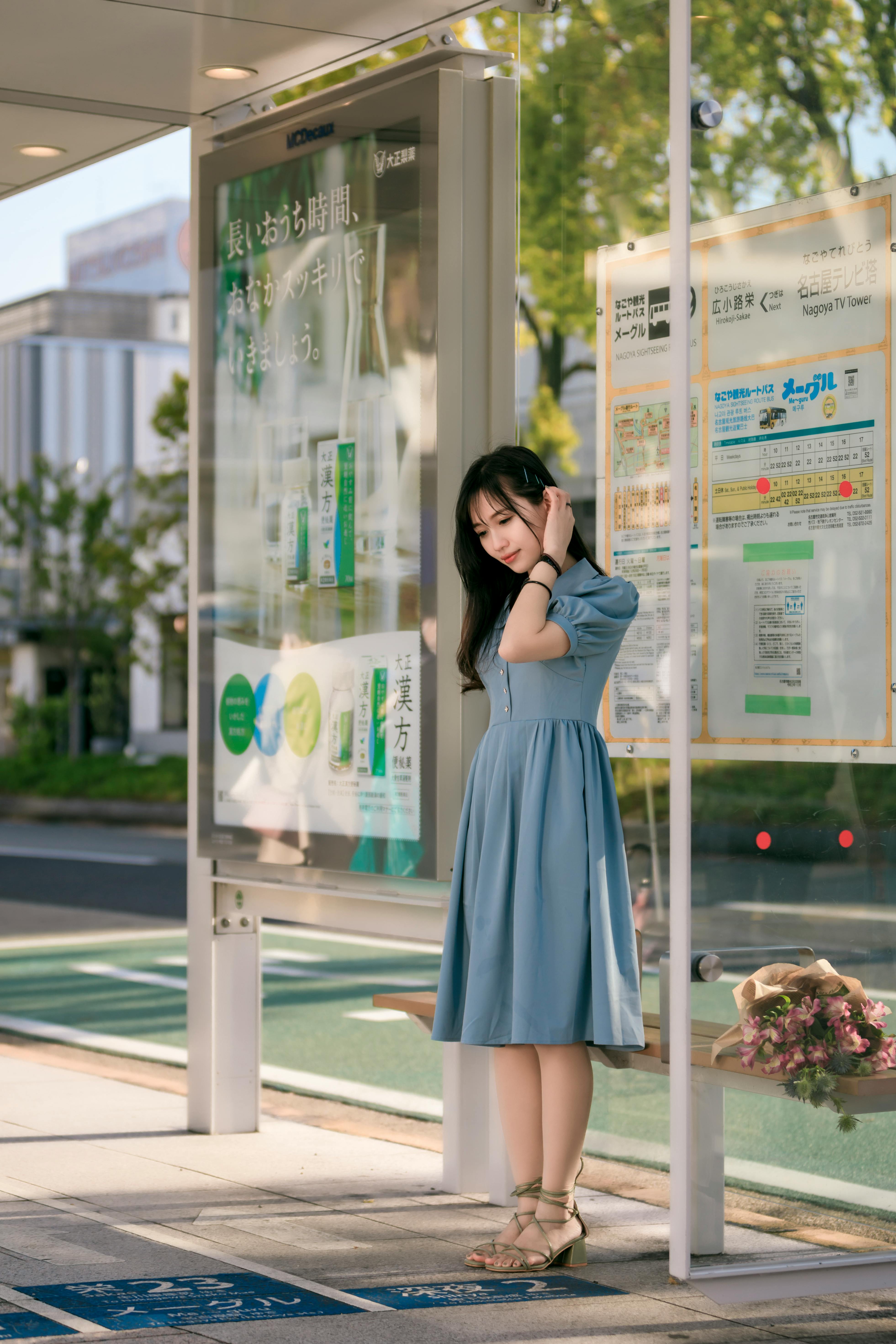 A woman in a blue dress stands pensively at a bus stop on a sunny day.