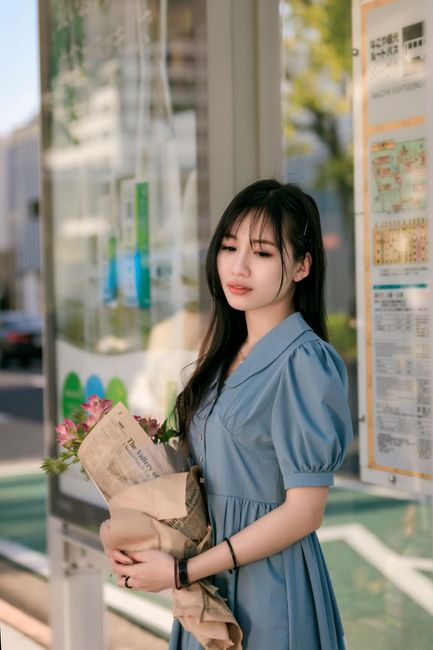 Woman in a blue dress with flowers at a bus stop, showcasing a serene moment outdoors.