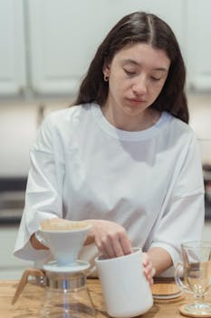 Young woman brewing coffee using a ceramic dripper in a cozy kitchen setting.