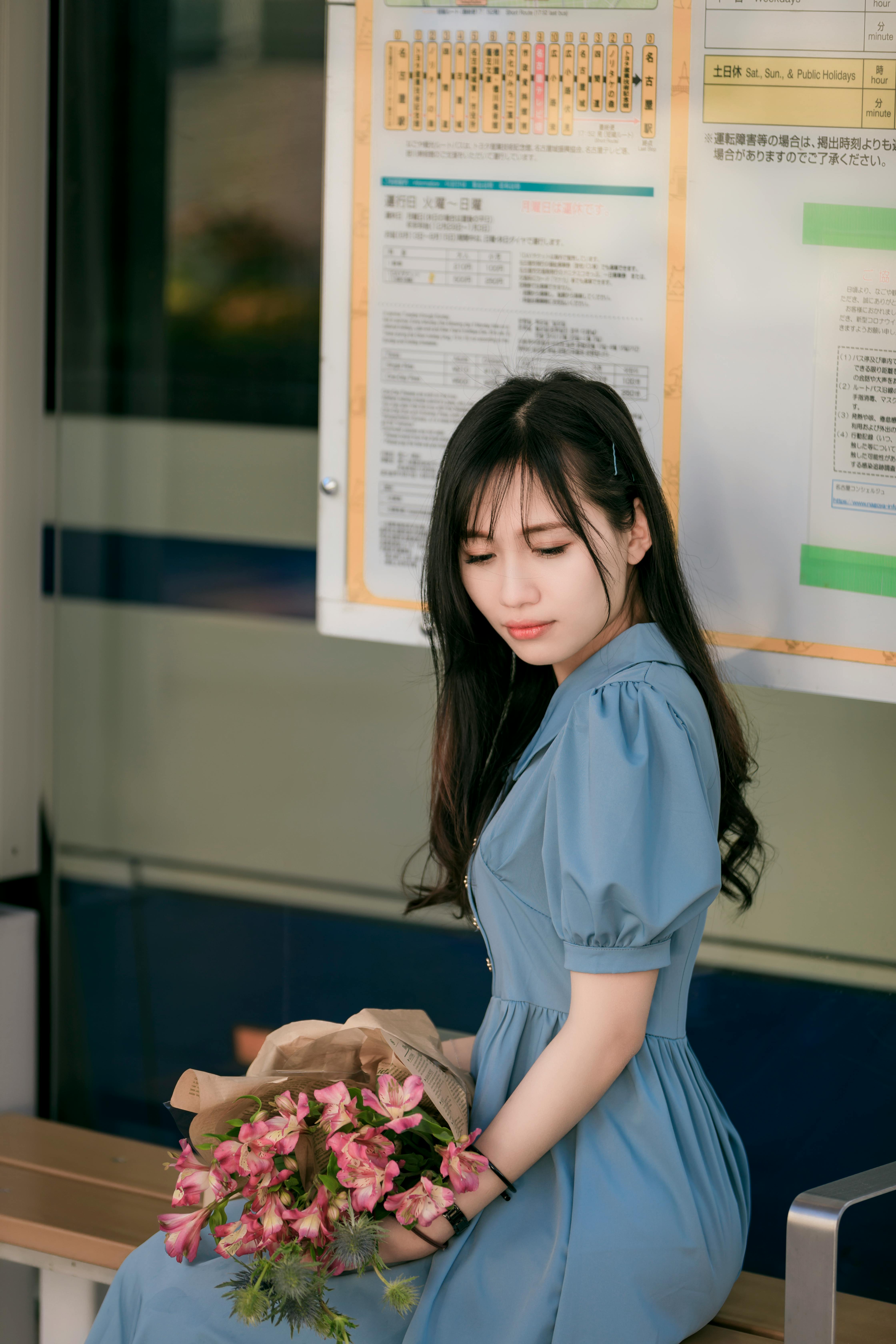 A thoughtful woman sitting indoors in a blue dress, holding a bouquet of flowers.
