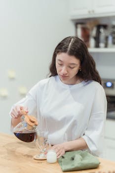 A woman in a white shirt pours coffee into a glass in a bright kitchen setting.