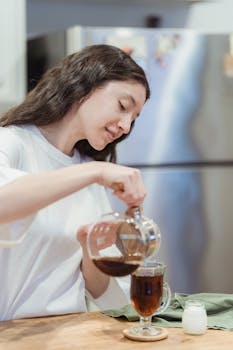 Young woman pours fresh coffee into a glass mug in her kitchen, enjoying a relaxing moment.