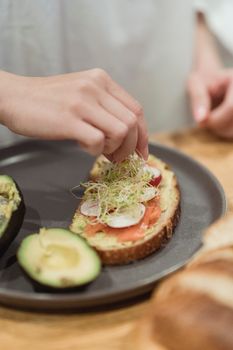 Close-up of hand preparing avocado toast with fresh vegetables on a plate, ideal for healthy breakfast concept.