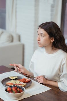 A woman in a white shirt enjoys a healthy breakfast indoors, featuring fruits and toast.