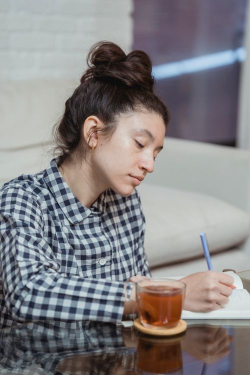 Free A young woman with long hair writing in a notebook while sitting indoors and drinking from a glass of tea. Stock Photo