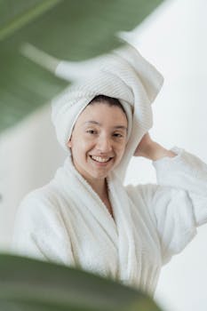 A cheerful woman wearing a white robe and towel turban smiling through tropical leaves indoors.