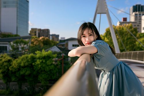Free Young woman in blue dress leaning on bridge railing with cityscape background. Stock Photo