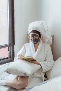 Woman enjoying a spa day at home, wearing a facial mask and reading a book in a cozy bedroom setting.