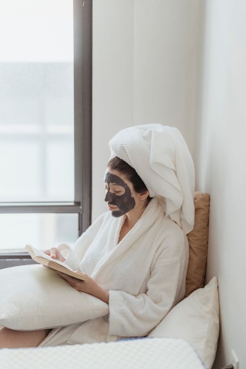 Free Woman in bathrobe with face mask reading by a window, symbolizing relaxation and self-care. Stock Photo
