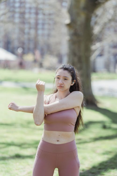 Free A woman in activewear stretching her arms in a sunny park setting, promoting outdoor fitness. Stock Photo