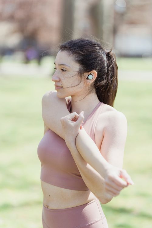 Free Young woman stretching her arms in a city park wearing fitness attire and wireless earbuds on a sunny day. Stock Photo