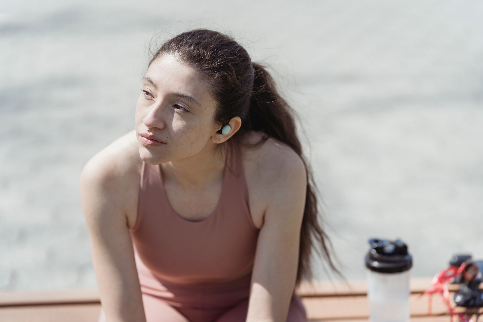 A young woman in sportswear relaxes outdoors with earbuds and a water bottle, enjoying leisure time.