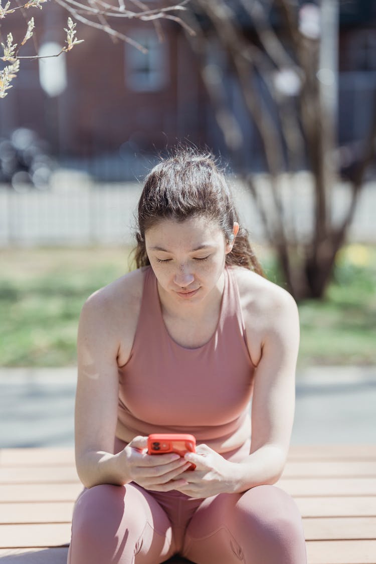 Woman In Sports Clothing Sitting Outdoors And Using Phone 