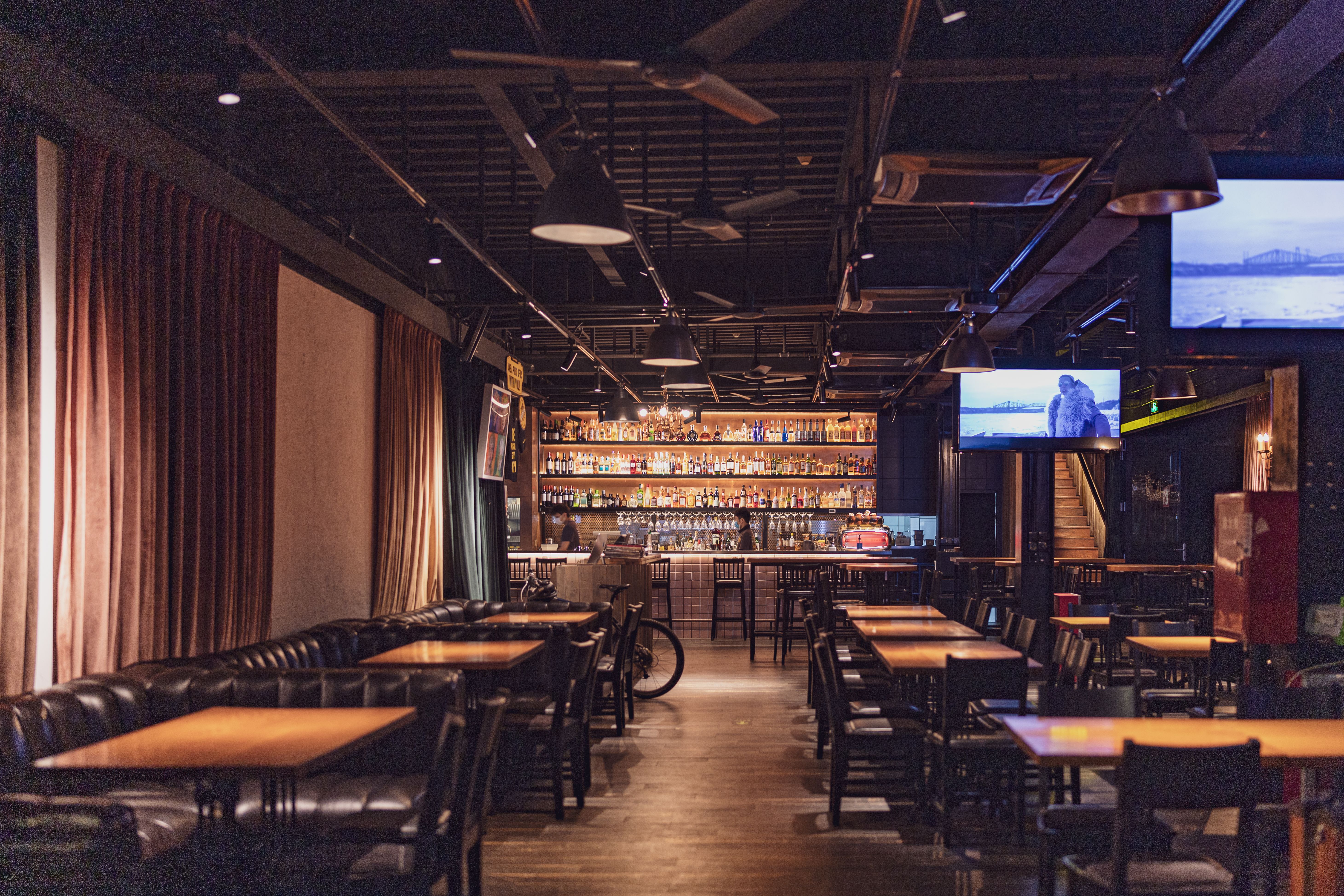 A spacious modern bar interior with wooden tables and chairs, ambient lighting, and a stocked bar.