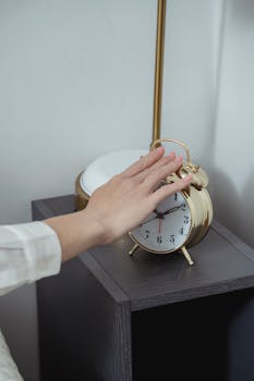 A hand reaching to turn off a vintage alarm clock on a bedside table.