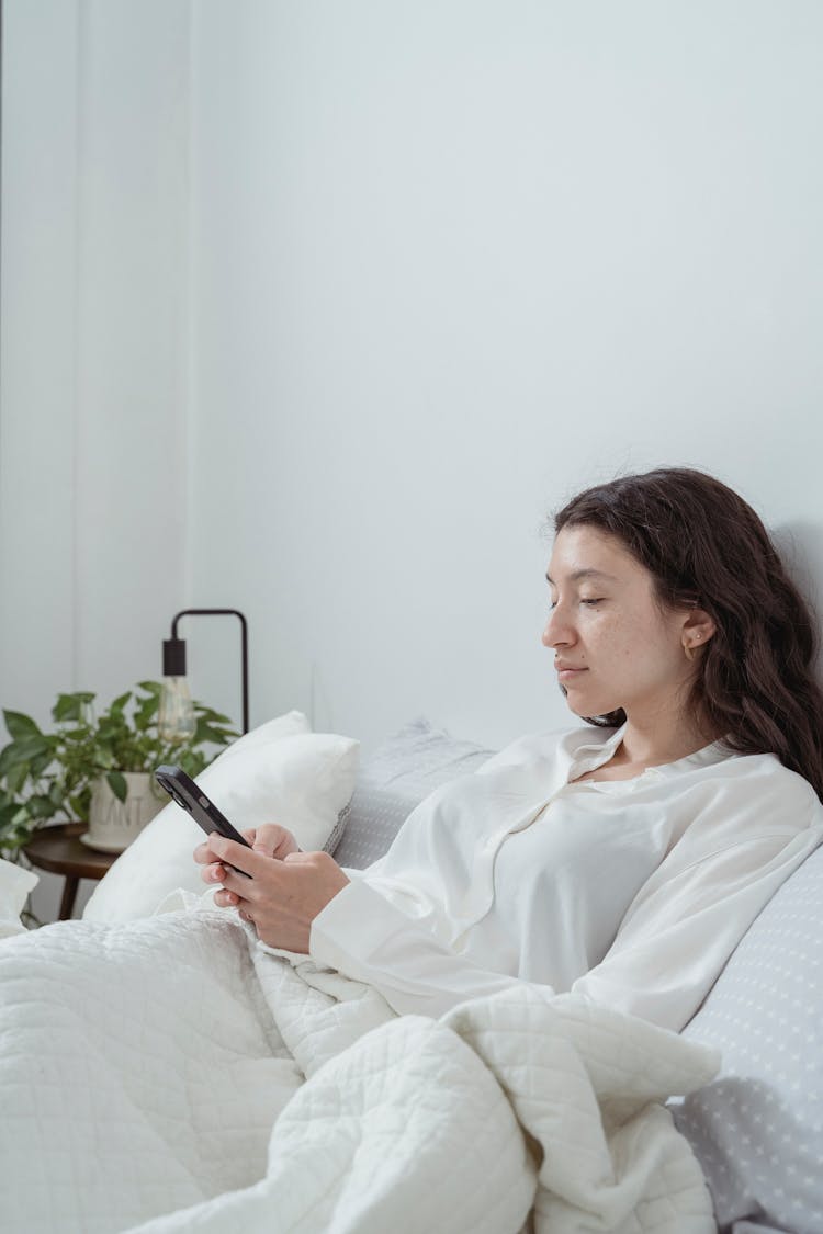 Woman Wearing White Pyjama Lying In Bed With Mobile Phone