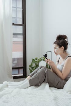 Woman with facial mask using smartphone while relaxing in a minimal bedroom setting.