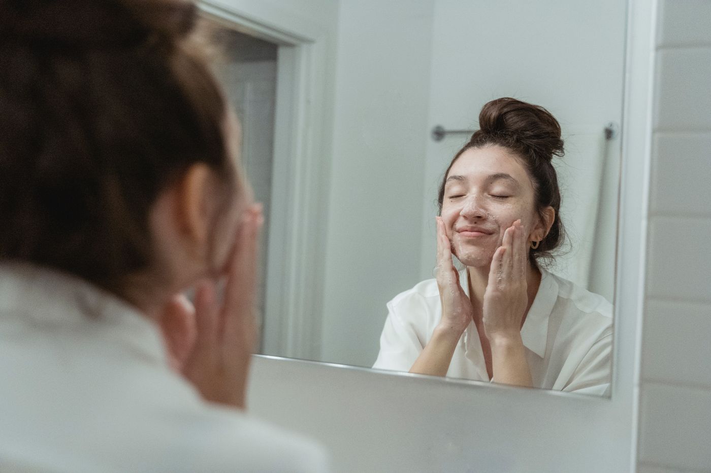 Woman cleansing her face with beauty care products beside a bathroom mirror