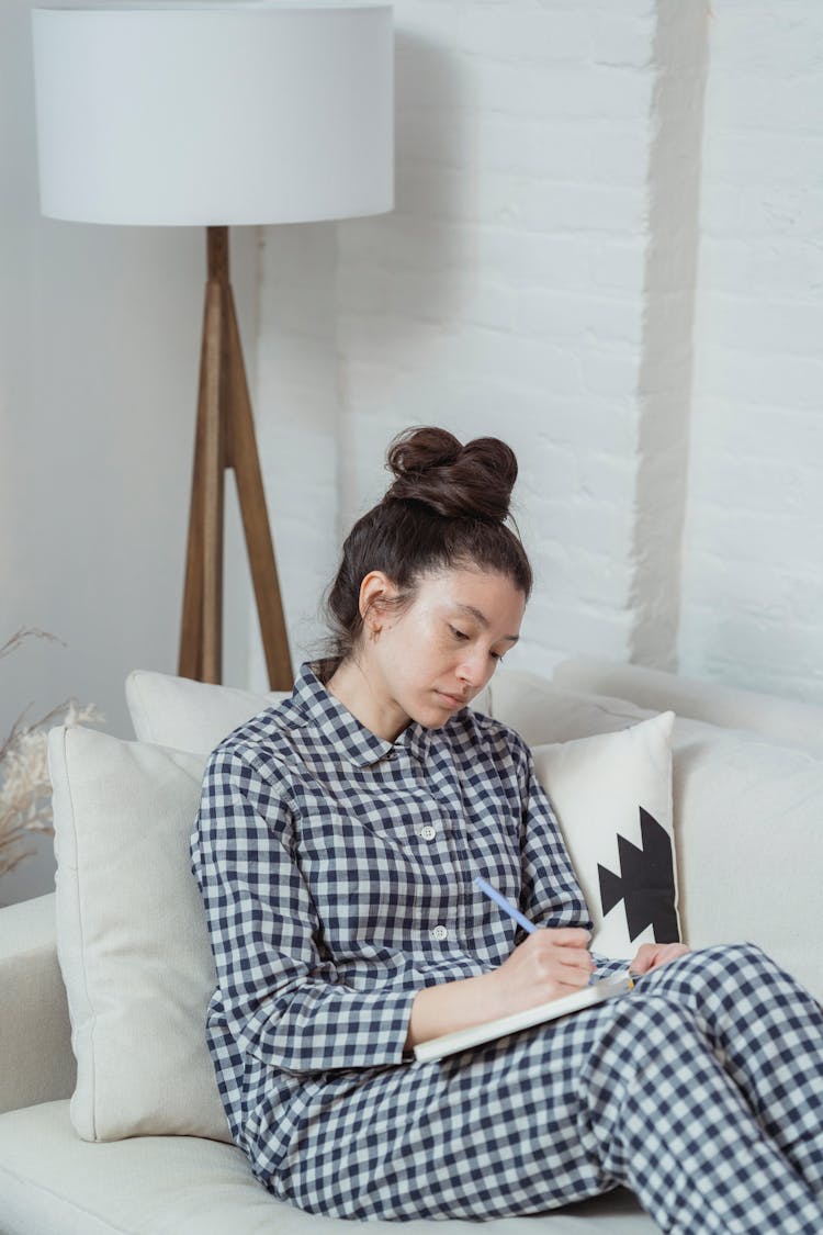 A Woman In Checkered Pajamas Sitting On The Couch While Writing On Notebook