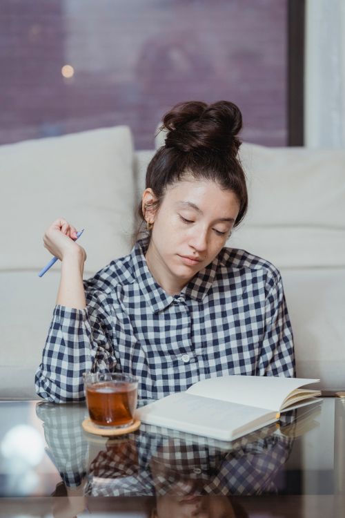 Free Young woman in plaid shirt reading a book indoors with a glass of tea on the table. Stock Photo
