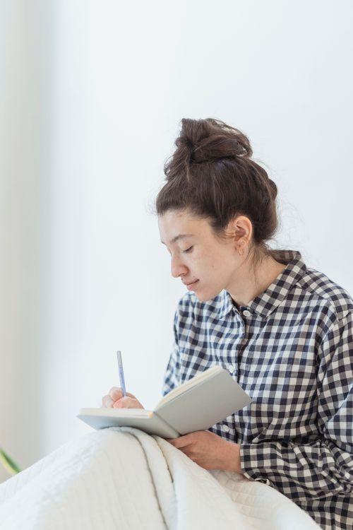 Free Young woman in plaid shirt writing in a notebook indoors, showcasing creativity and concentration. Stock Photo