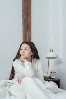 Woman in pajamas sitting thoughtfully in bed with a cozy blanket.