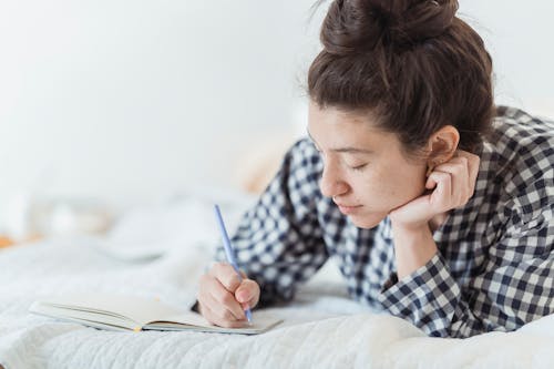 Free A Woman in Checkered Long Sleeves Writing on a Notebook while Lying on the Bed Stock Photo