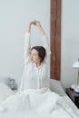 A Woman in White Sleepwear Stretching Her Arms while Sitting on the Bed