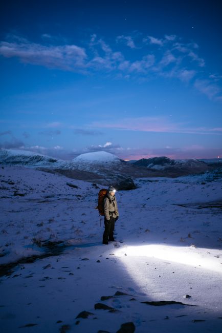 Hiker with headlamp trekking through snowy terrain at twilight in Capel Curig, Wales.