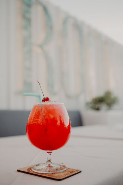 Glass of delicious red cocktail with ice and cherry on top placed on coaster on white table on veranda of restaurant in evening