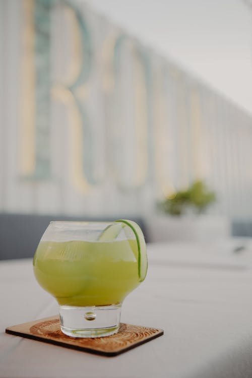 Free Glass of refreshing cold yellow cocktail with cucumber placed on coaster on white table in veranda of restaurant Stock Photo