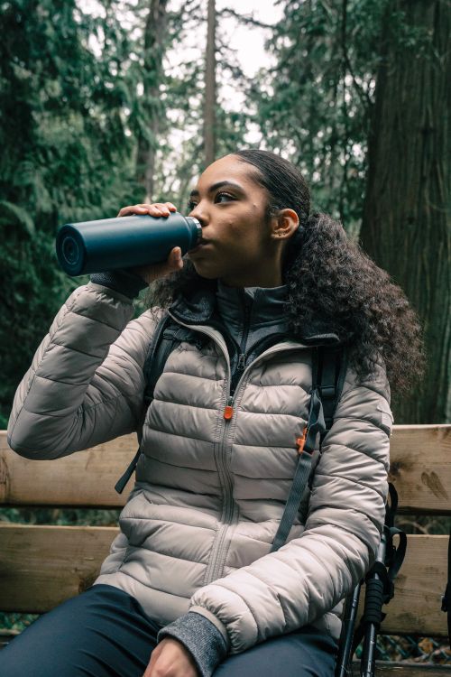 Free A woman sitting on a bench in a forest drinking from a tumbler, dressed in hiking gear. Stock Photo