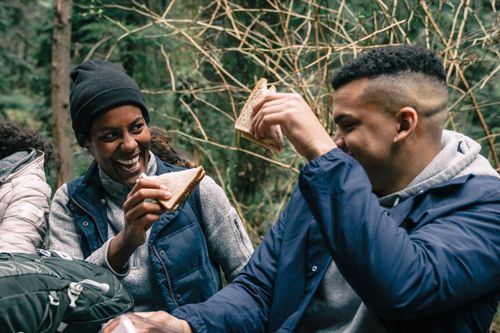 Free Two friends laughing and sharing sandwiches during an outdoor picnic in the woods. Stock Photo