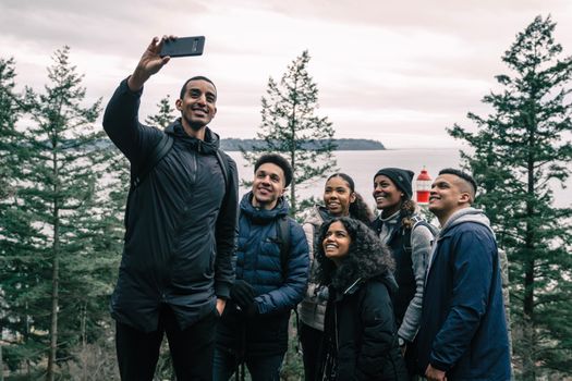 A diverse group of friends enjoying a hiking adventure and taking a selfie outdoors near a scenic lighthouse.