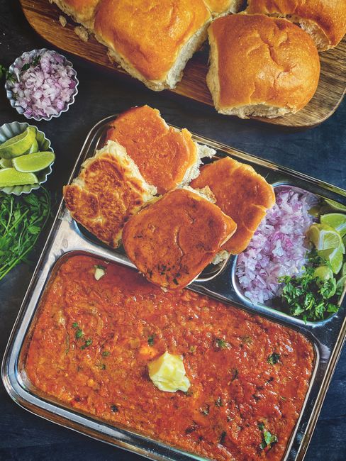 A wide skillet of thick, orange-red mashed vegetable pav bhaji topped with a pat of melting butter, chopped onion, and cilantro, next to a plate of buttered, griddle-toasted pav buns.