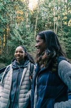 Two women standing by a car, smiling and laughing in a forest setting, dressed warmly for fall.