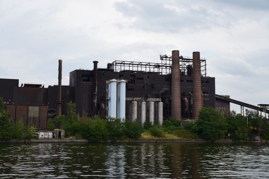 A large industrial factory with chimneys by a river, surrounded by trees on a cloudy day.