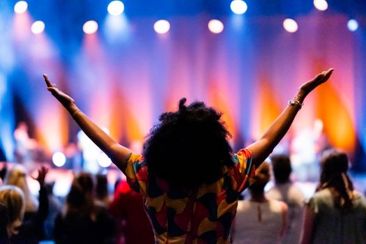 Back view of anonymous ethnic female viewer enjoying performance with arms apart against people and stage with shiny spotlights