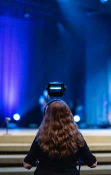 Back view of anonymous woman in headset recording video of live performance on camera against stage