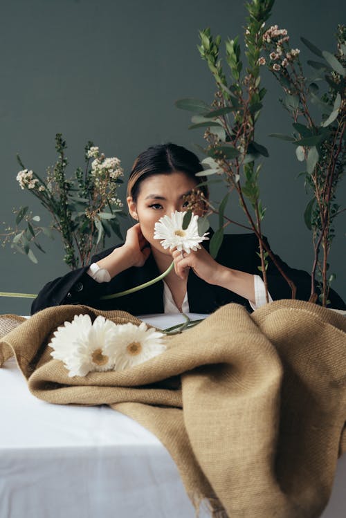 Confident ethnic female smelling white blooming gerbera flower at table with floral branches and sackcloth and looking at camera