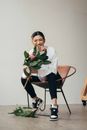 Happy young Asian female in stylish clothes sitting on modern armchair while laughing after dropping flower in light studio
