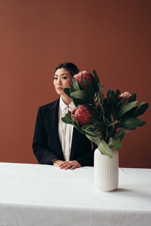 Elegant female in formal outfit sitting at white table with flowers in vase while looking at camera on brown background