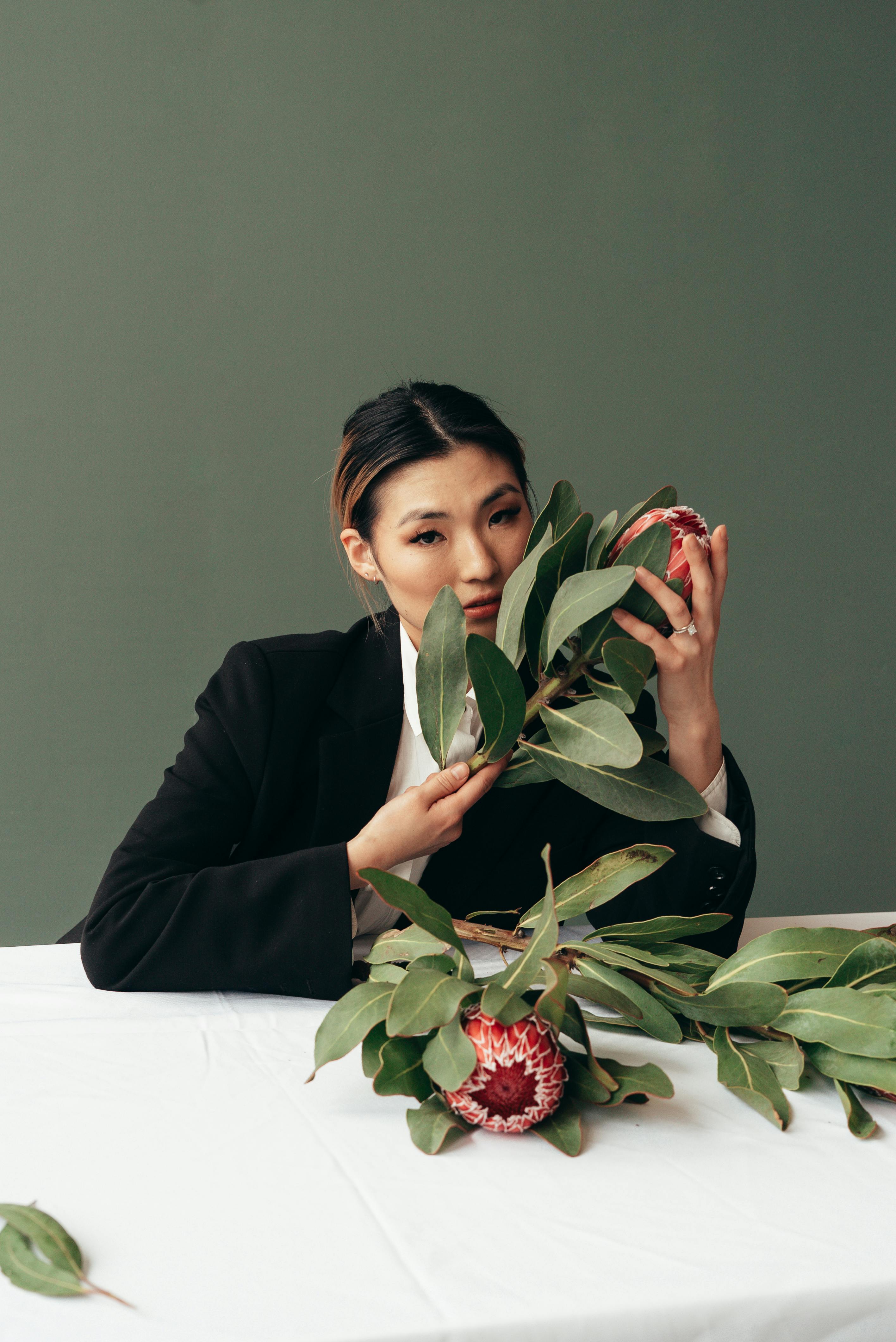 Ethnic female with blooming pink Protea flowers in black jacket looking at camera on gray background