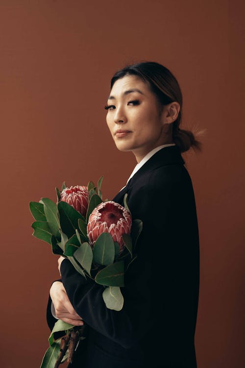 Calm ethnic female with fresh delicate Protea flowers in blossom looking at camera on red background of studio